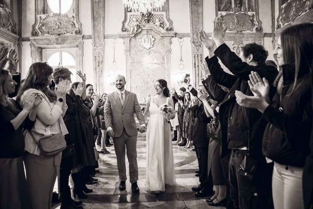 Bride and groom walk down the aisle as guests applaud in an elegant historic venue.