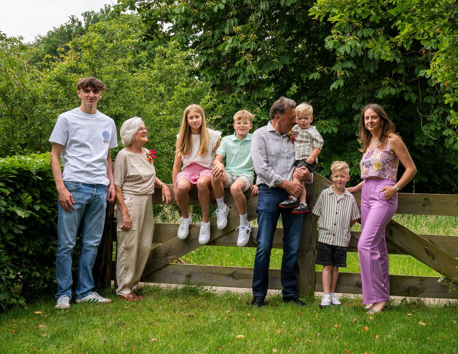 Een grote familiegroep poseert buiten bij een houten hek, omringd door weelderige groene bomen, op een zonnige dag.