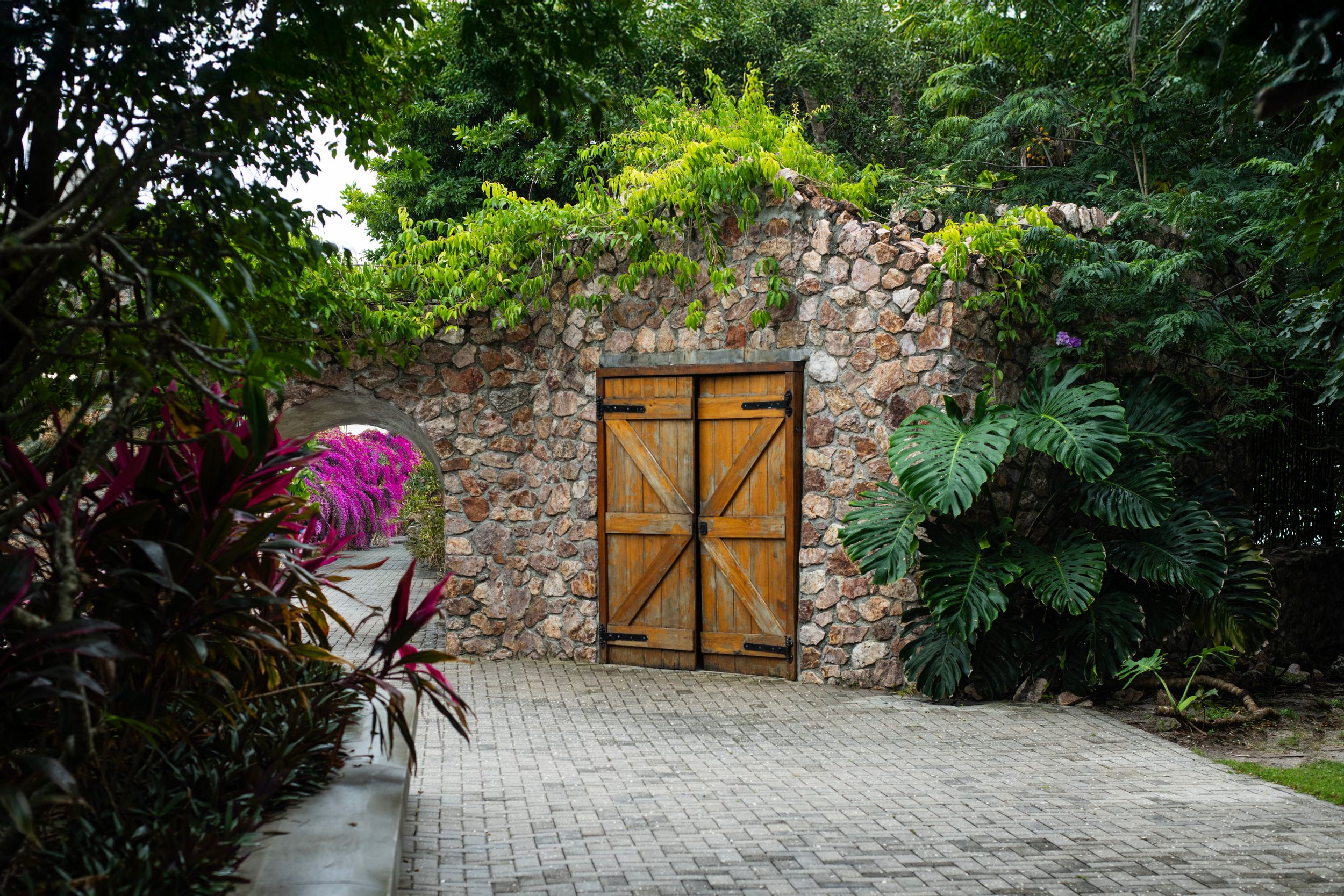 Wooden double doors set in a rustic stone wall surrounded by lush tropical greenery and pink flowers on a paved courtyard.