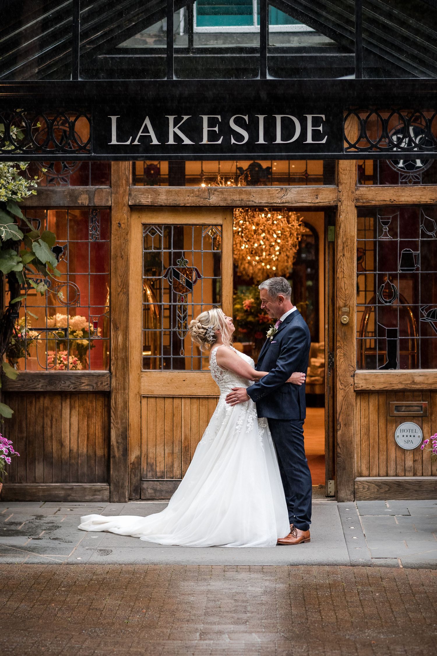 Bride and groom embracing outside Lakeside venue entrance with warm glowing interior and wooden doors.