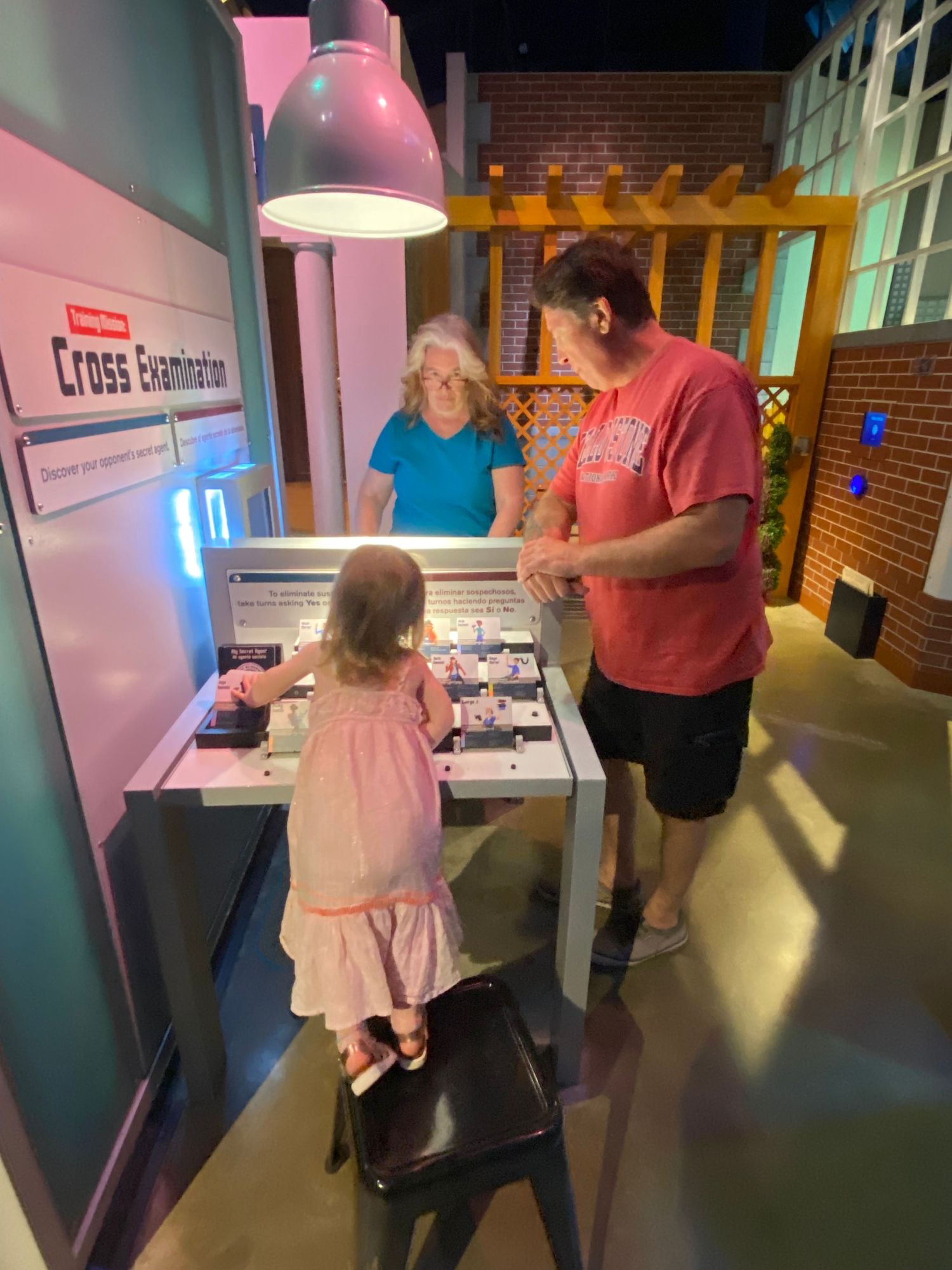 Family exploring Cross Examination interactive exhibit at children's museum, with toddler girl and adults at illuminated display table.