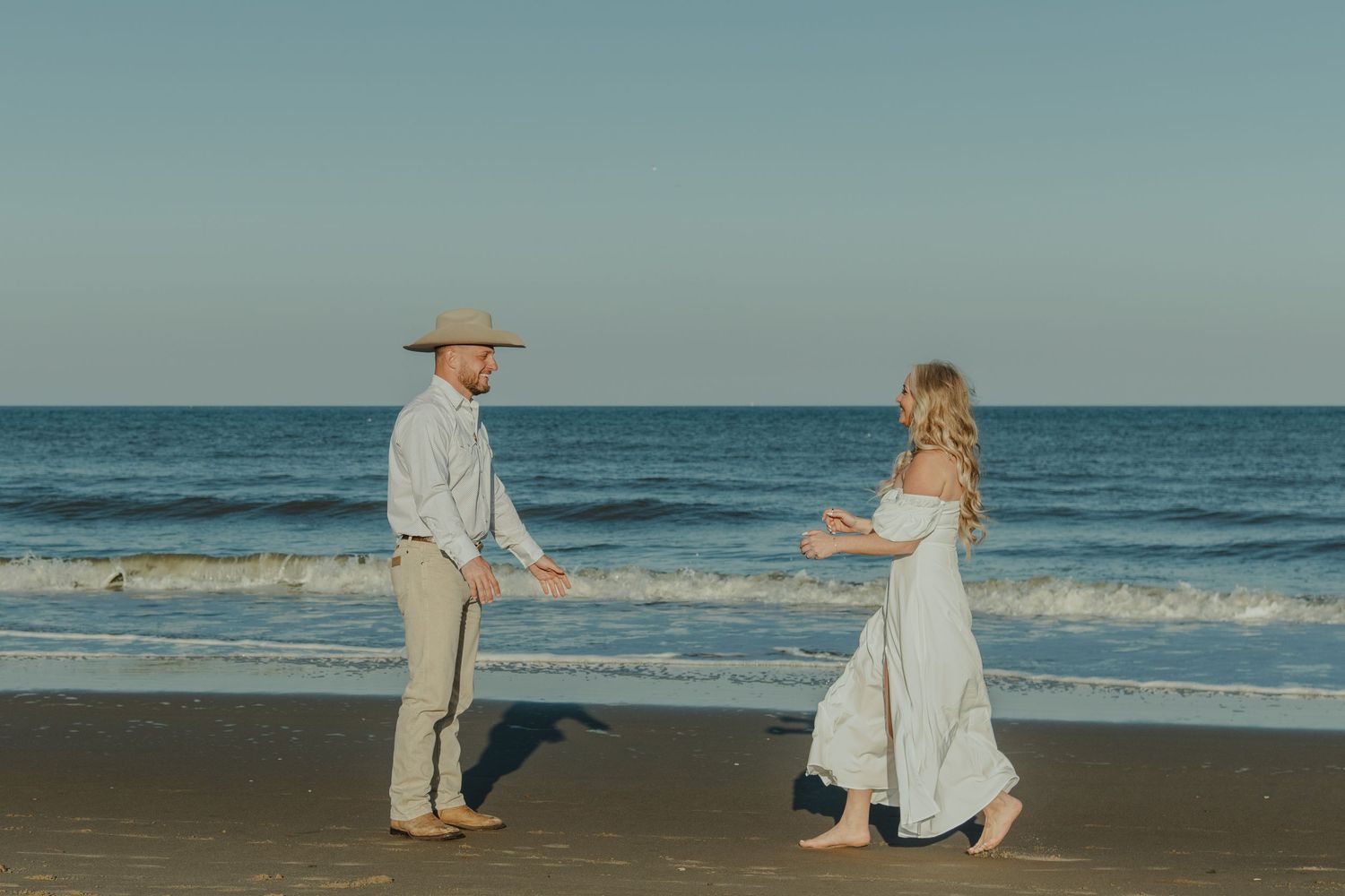 Couple in white outfits reaching toward each other on a sandy beach with ocean waves in the background.