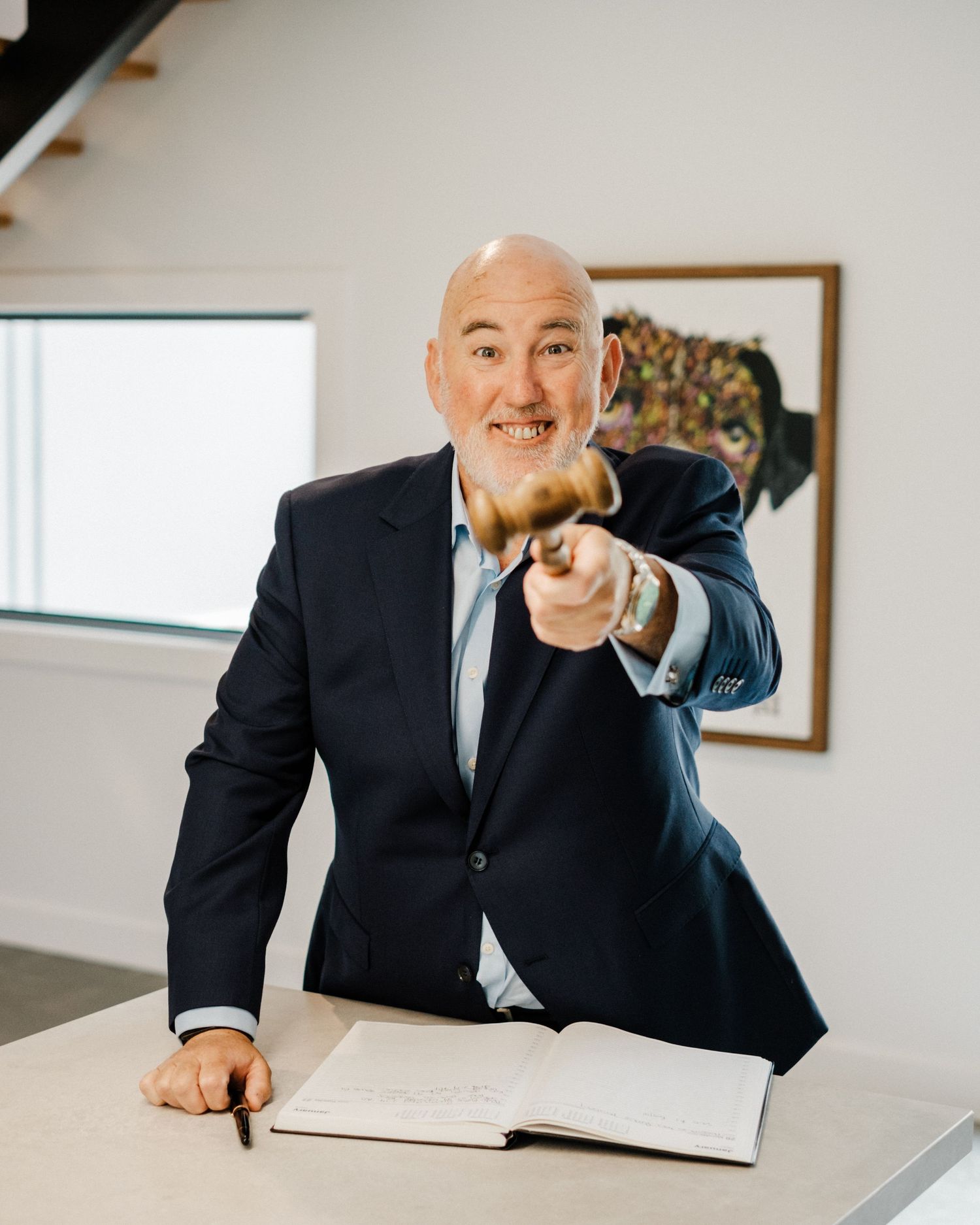 Bald man in navy suit leaning over desk pointing gavel toward camera with open book in bright office.