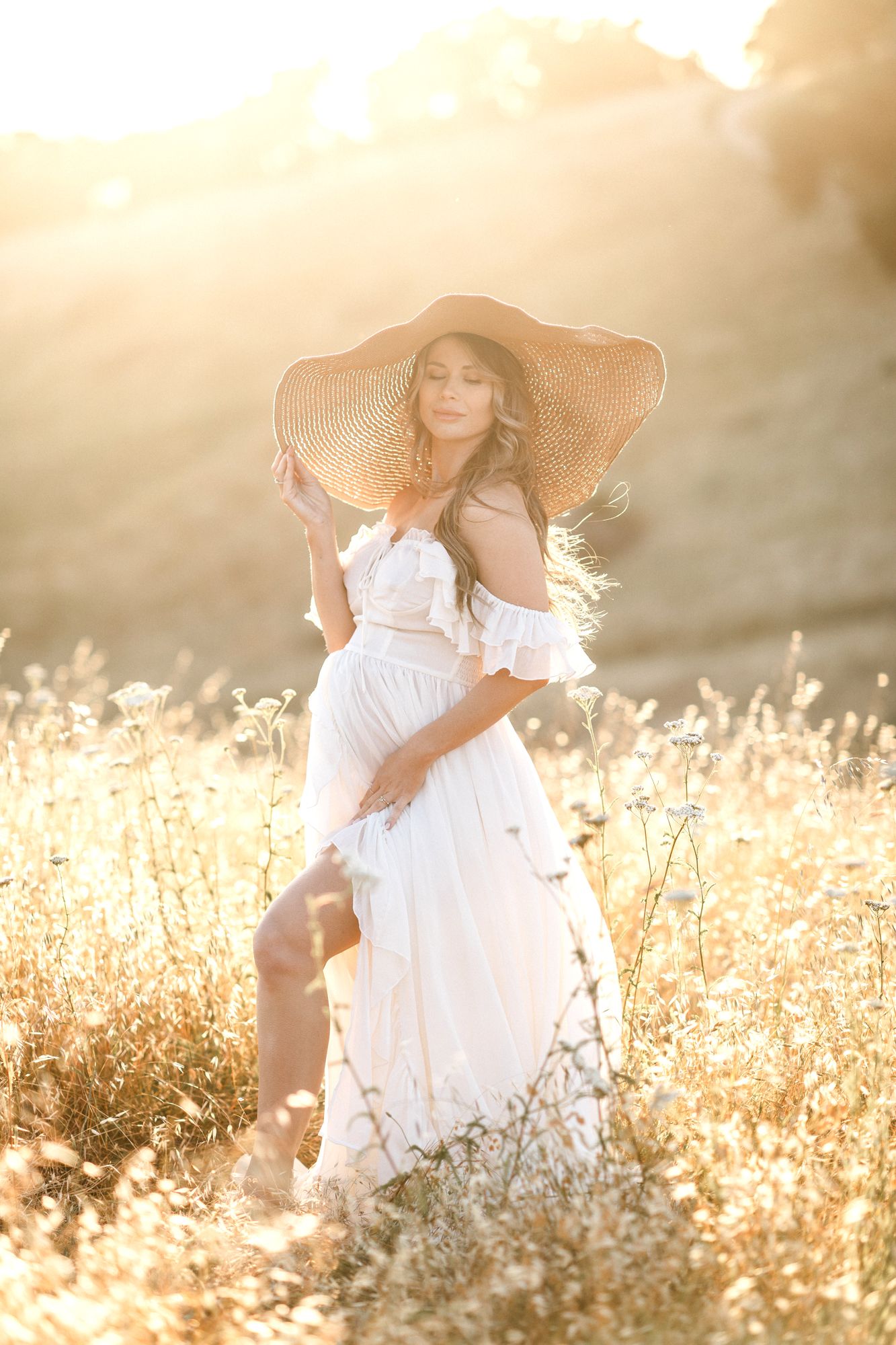 Expectant mother in sheer white maternity gown poses with a straw hat on in a golden field. Lakes Region NH maternity photographer