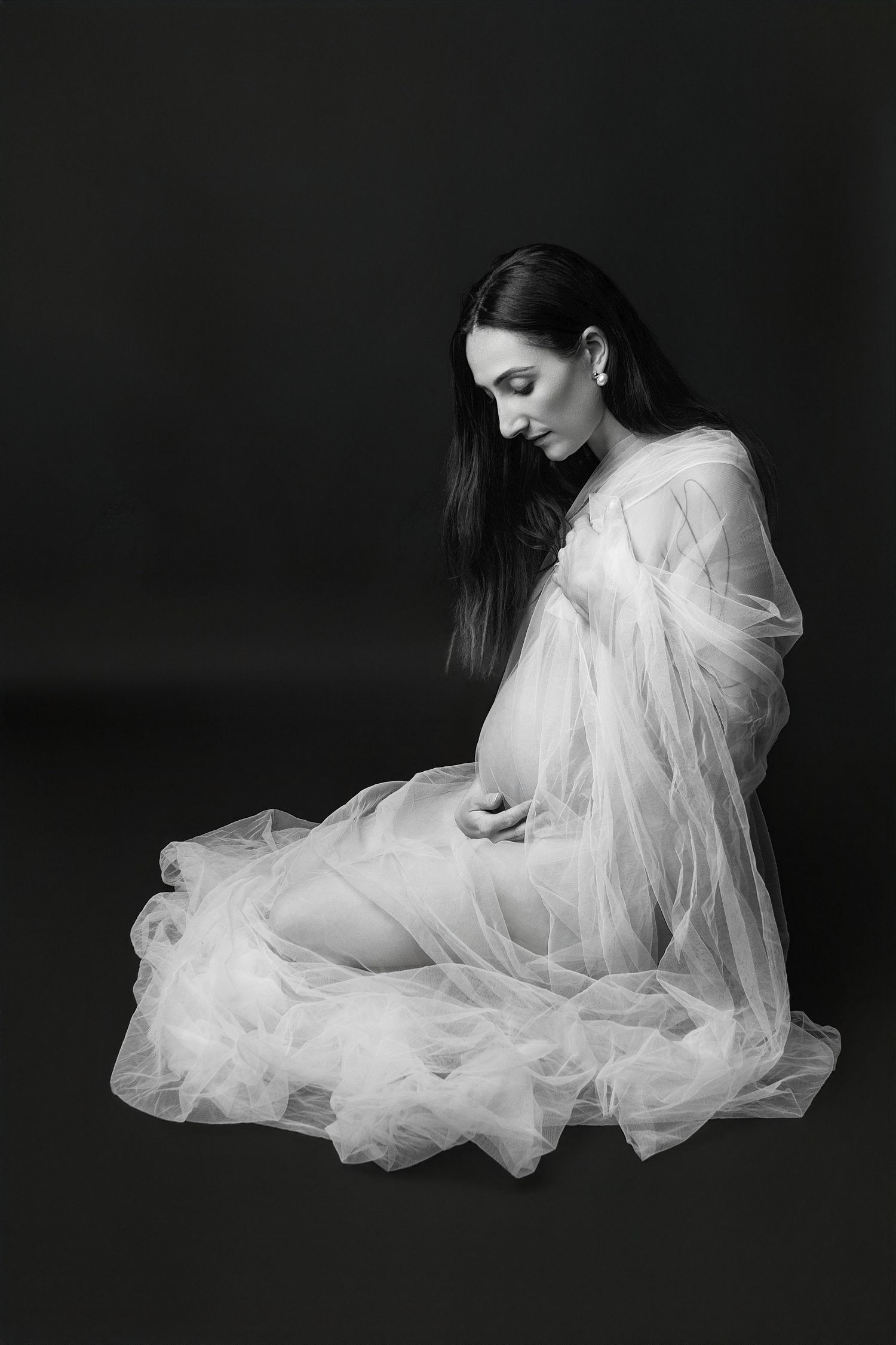 Black and white maternity photo of a pregnant woman in flowing white tulle gown sitting gracefully against dark background.