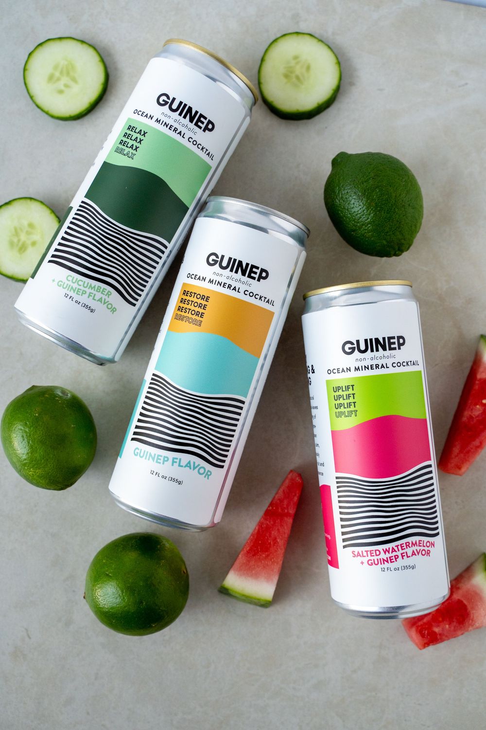 Colorful canned beverages from Guined brand arranged with fresh cucumber slices, limes and watermelon on light surface.