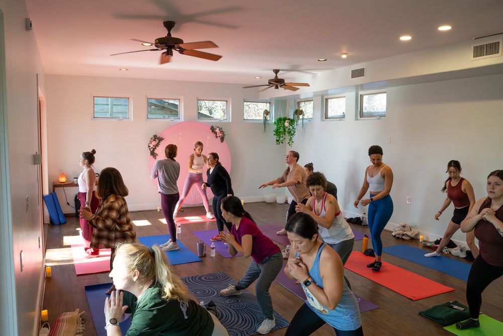 Group yoga class with participants practicing poses on colorful mats in a bright, modern studio with ceiling fans.