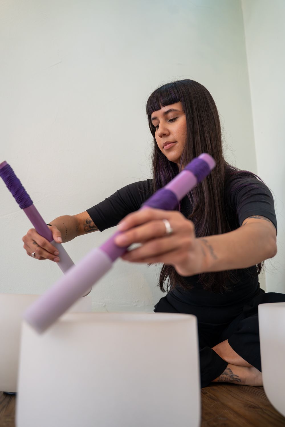 Woman in black outfit holding purple mallets near crystal singing bowls in a bright studio.