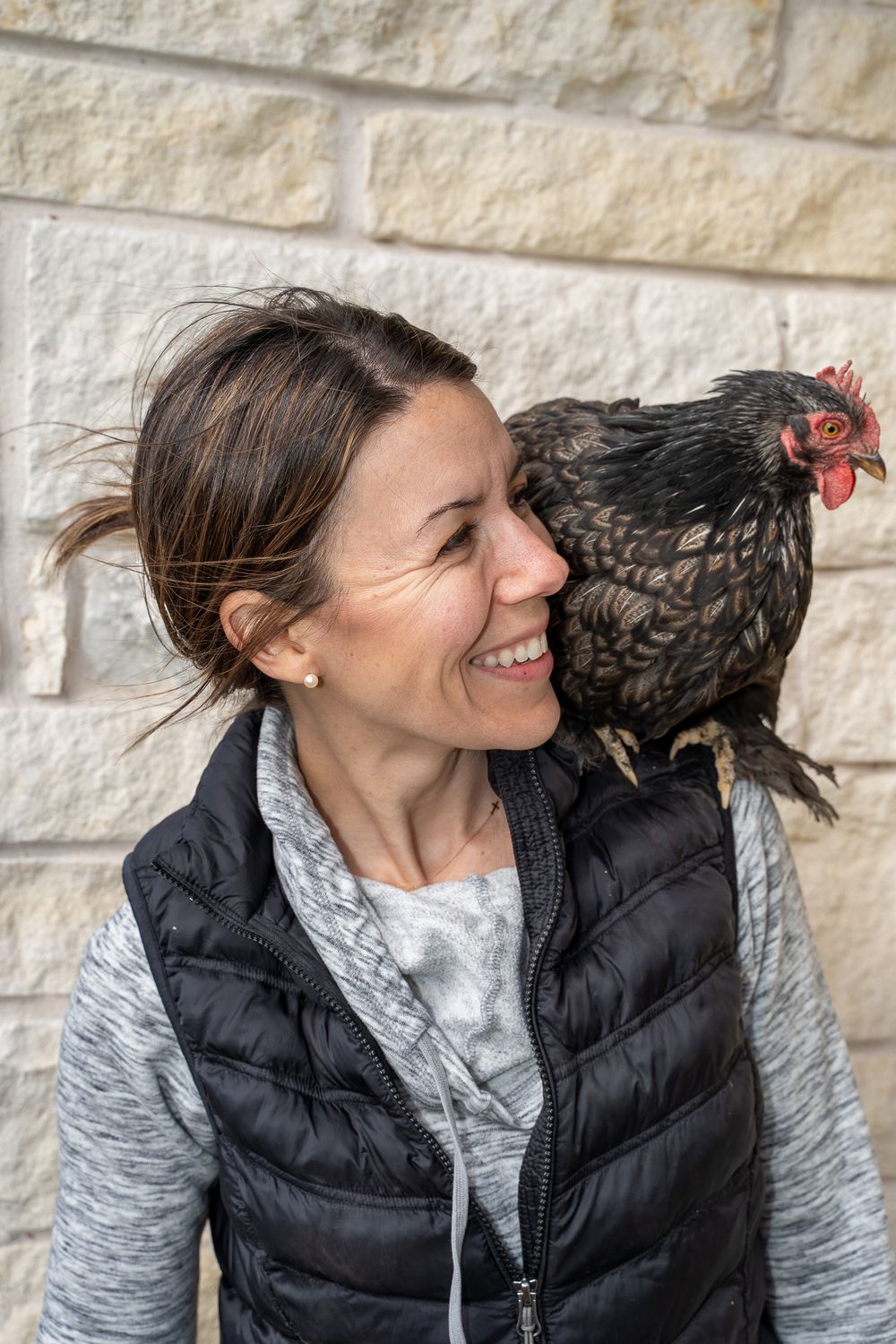 Smiling woman with a black hen perched on her shoulder against a stone wall background.