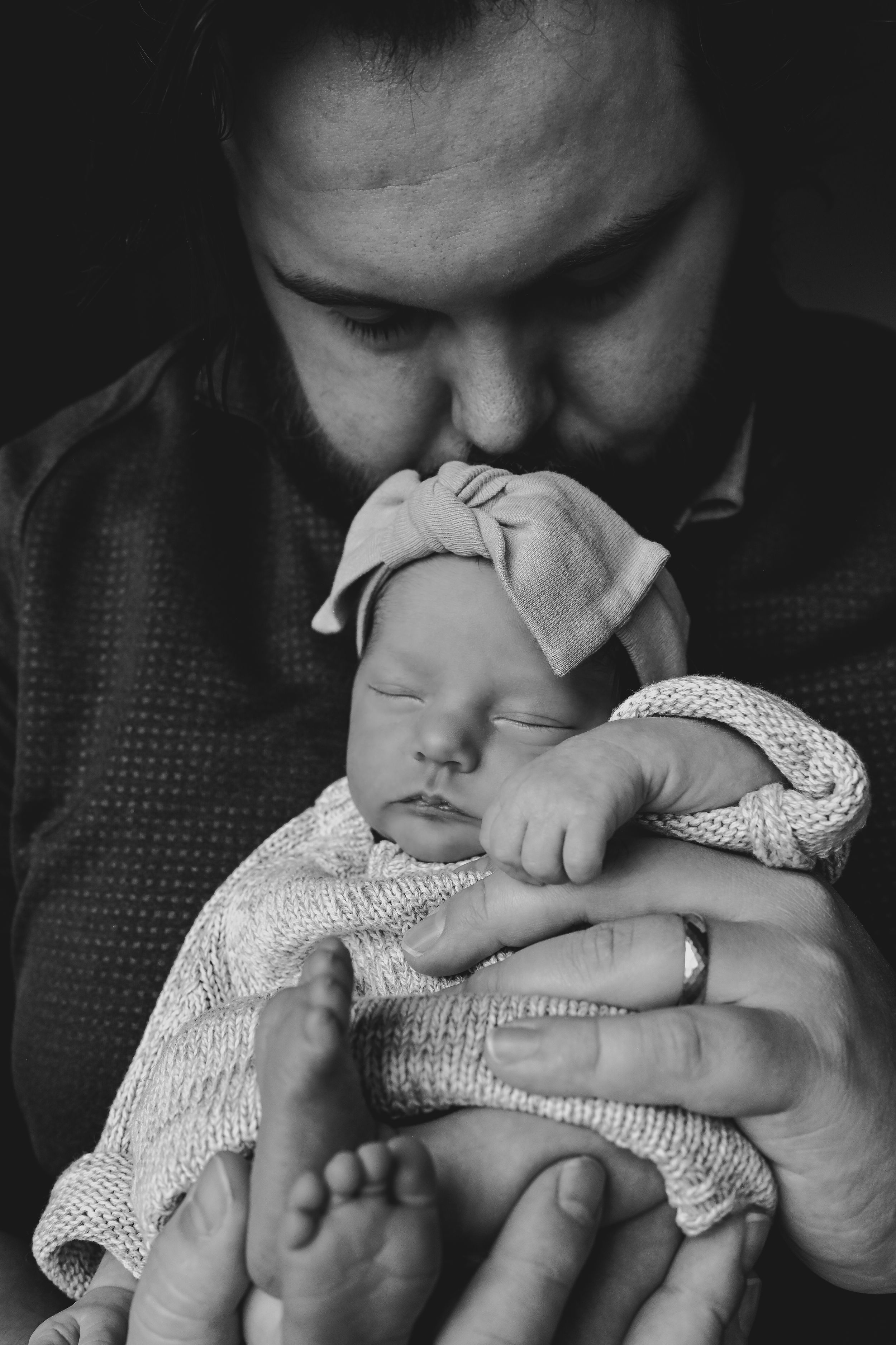 Black and white photo of a bearded father tenderly kissing his sleeping newborn baby girl wearing a bow headband.