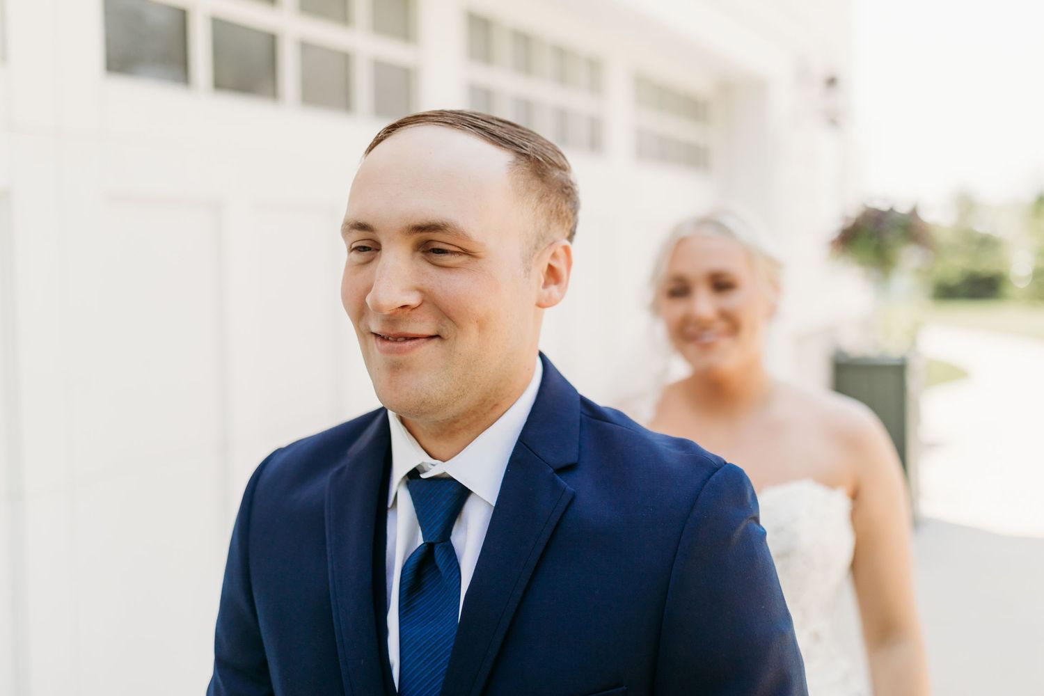 Groom in navy blue suit and tie smiling outdoors with bride in white wedding dress softly blurred in background.
