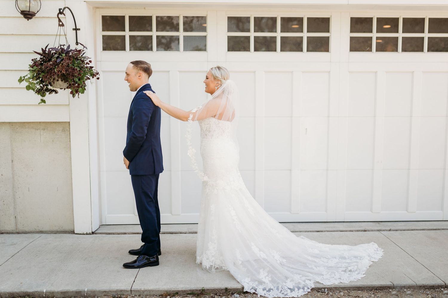 Bride in lace gown touches groom in navy suit during first look moment outside white garage doors.
