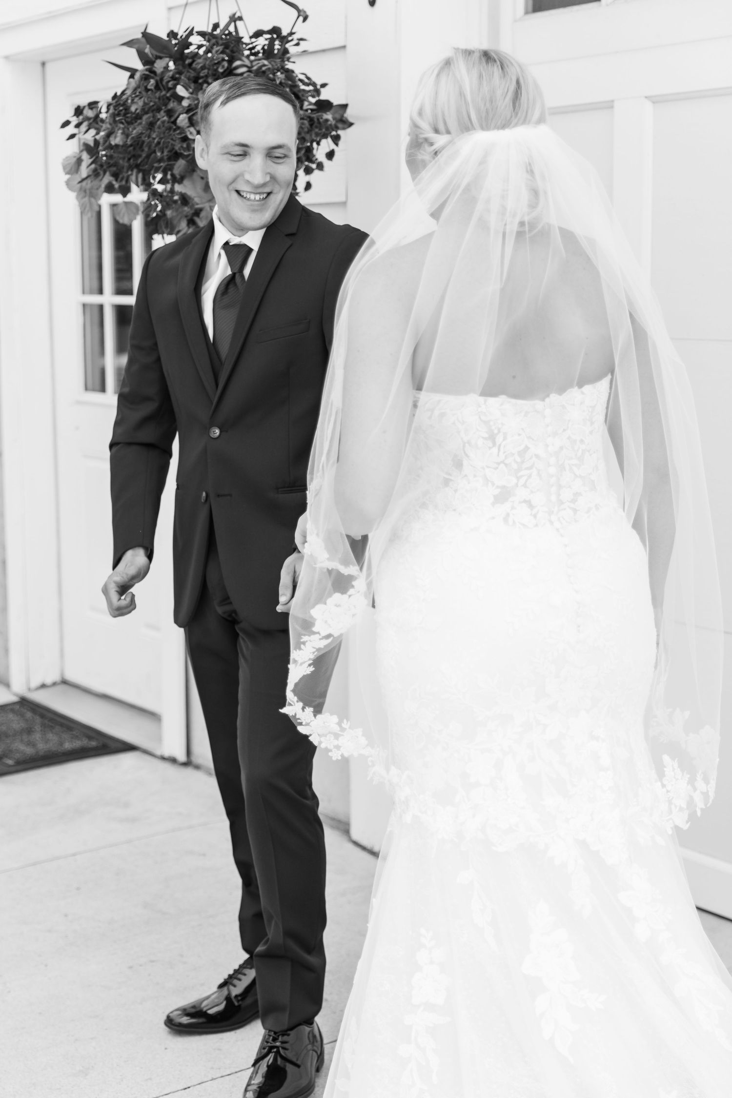 Black and white photo of a groom smiling at his bride during a first look outside a white building.
