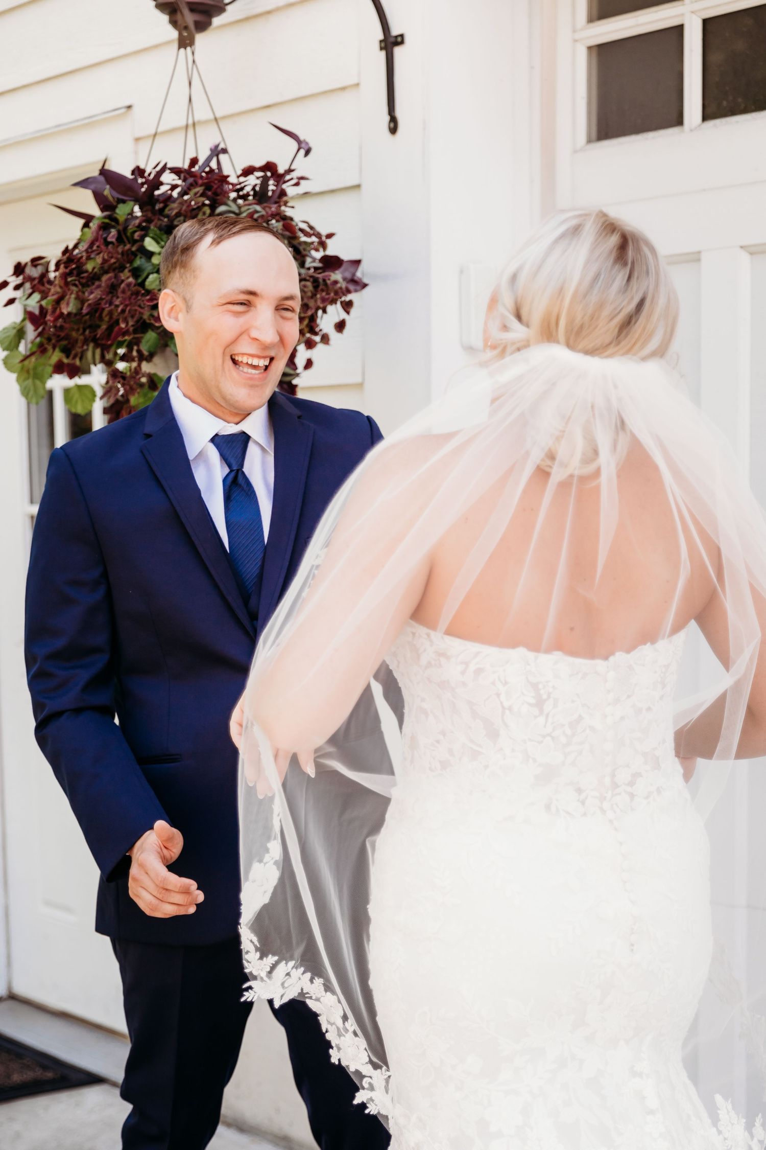 Groom in navy suit smiling at bride in lace wedding dress and veil during romantic first look moment.