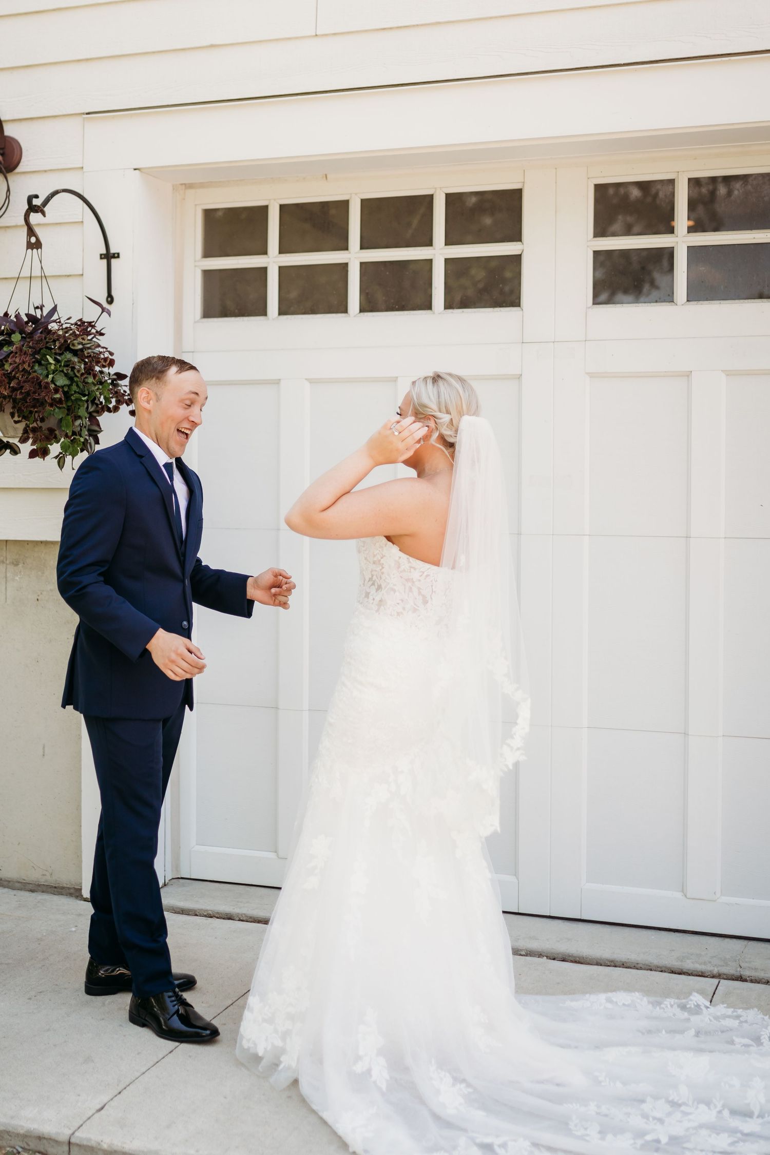 Bride in lace gown and veil shares emotional first look moment with groom in navy suit outside white garage doors.