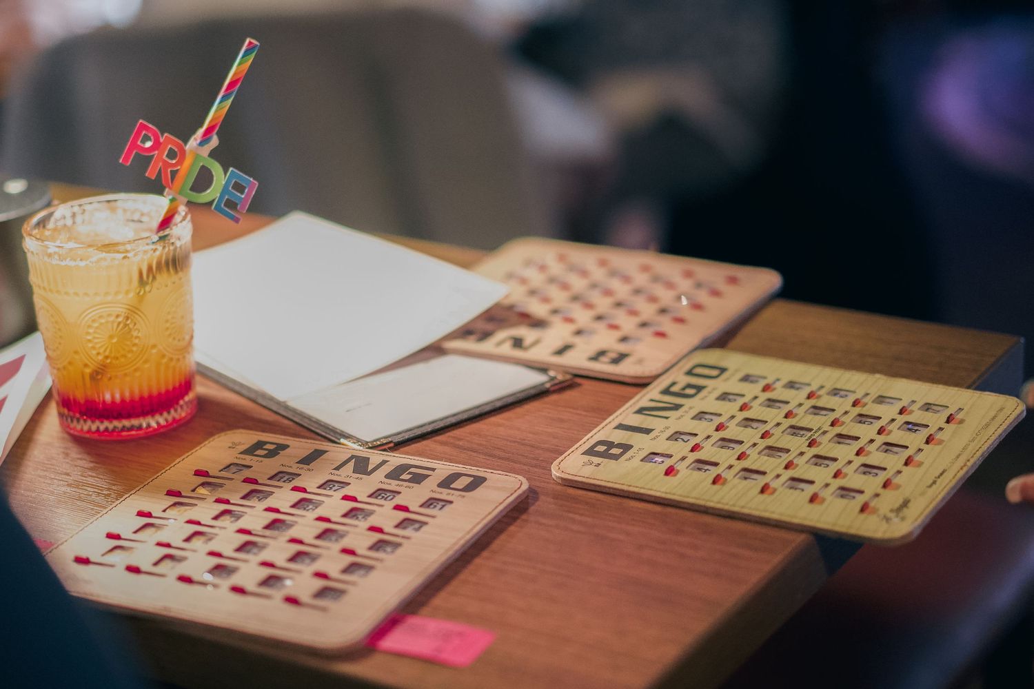 Bingo cards on a wooden table with a colorful cocktail featuring a Pride straw at a festive event.