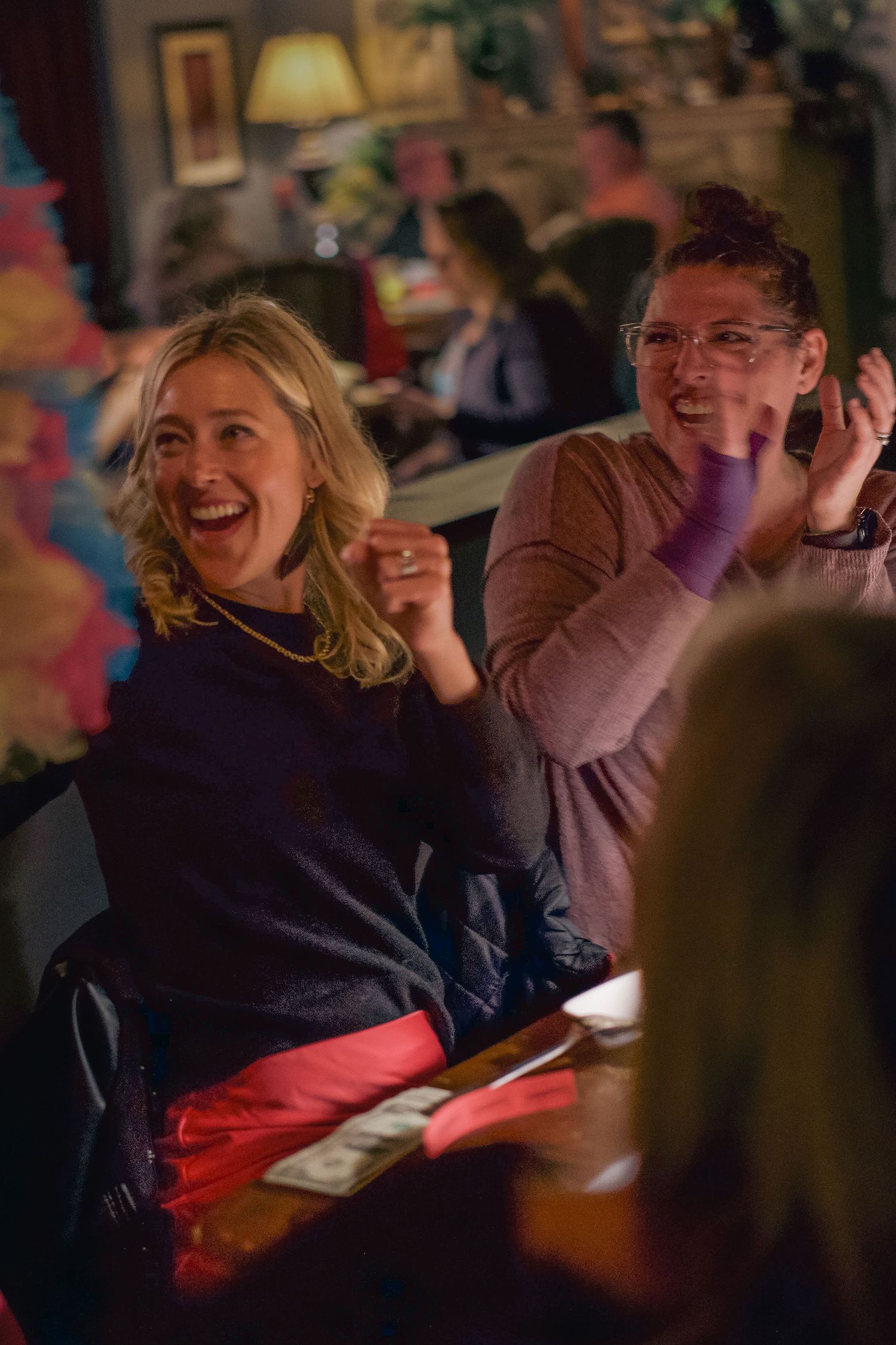 Two women laughing and clapping together at a restaurant table during a lively social gathering.