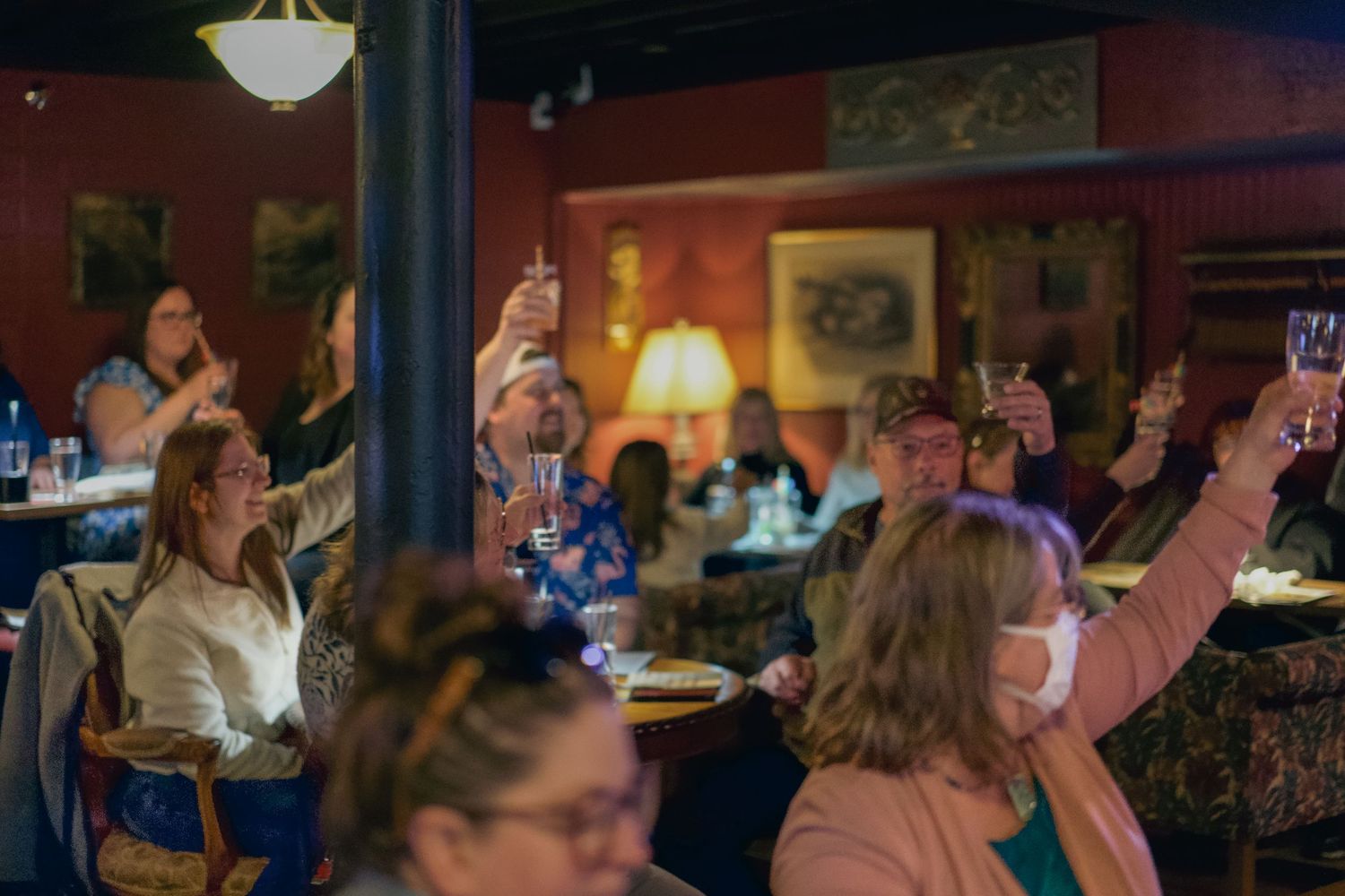 Group of people raising glasses in a toast at a dimly lit restaurant with red walls and warm lighting.