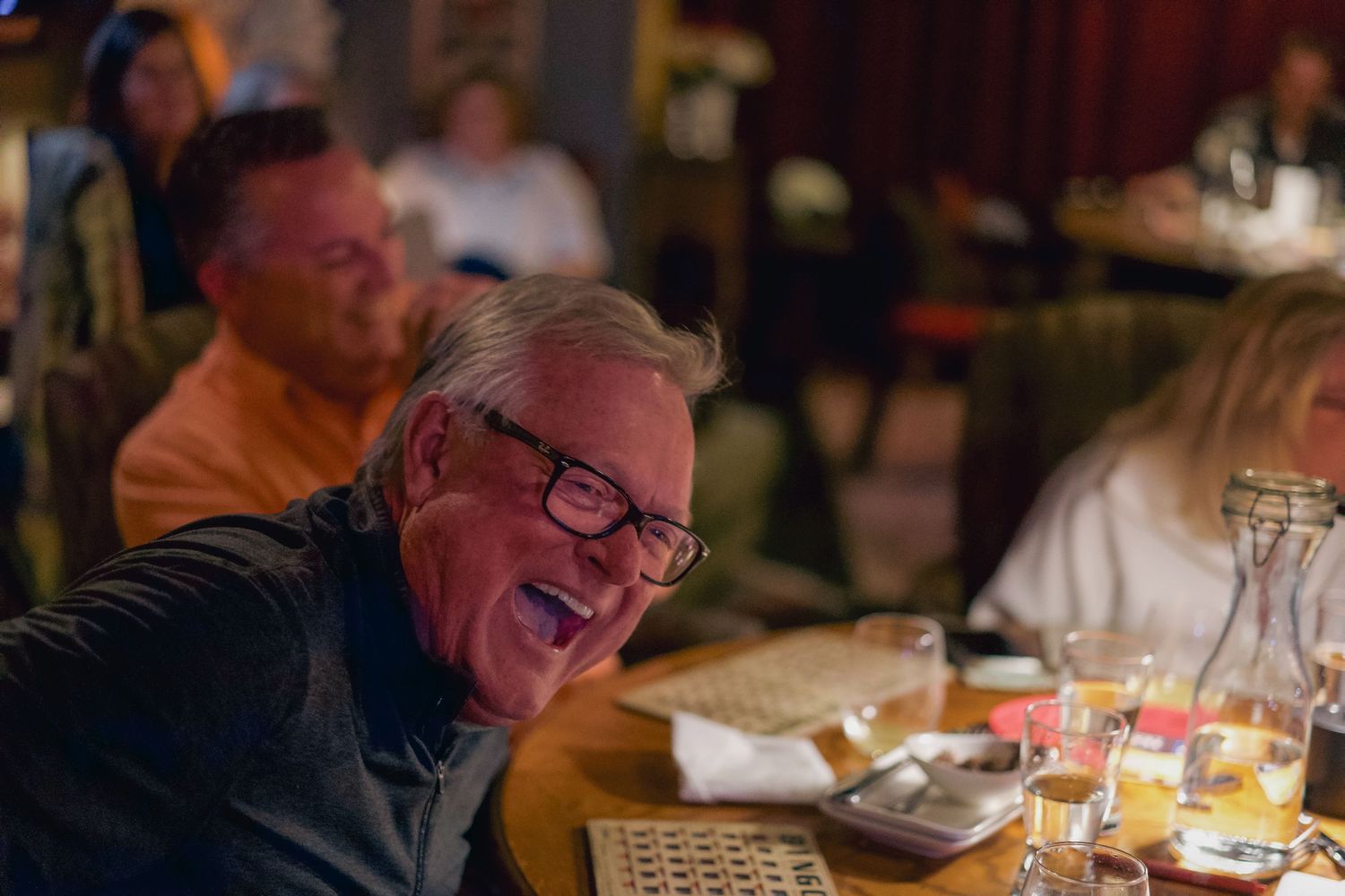Elderly man laughing joyfully at a restaurant dinner table with friends dining in a warmly lit setting.