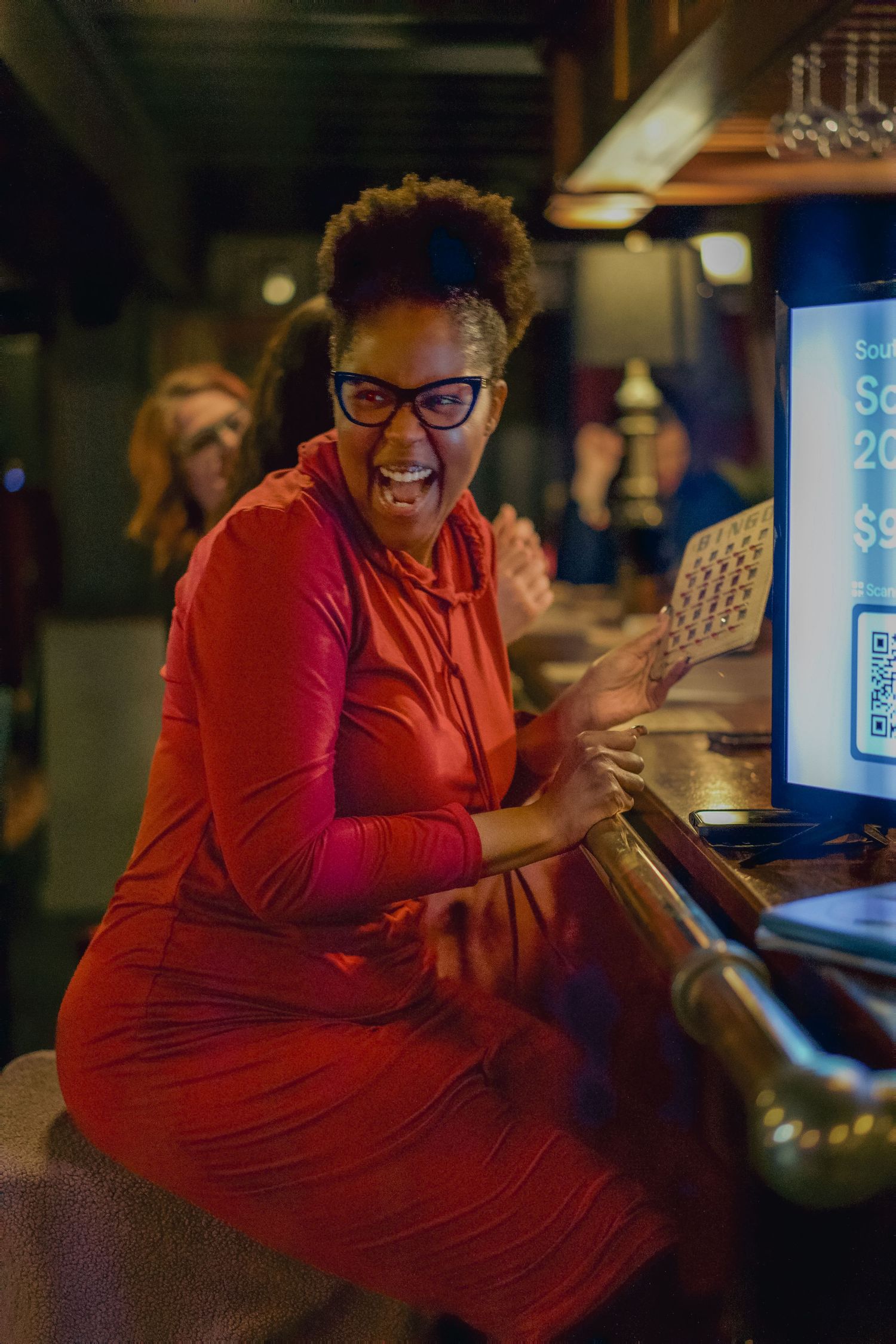 Excited woman in red dress holding paper at bar counter, wearing glasses, laughing with joy in dimly lit venue.