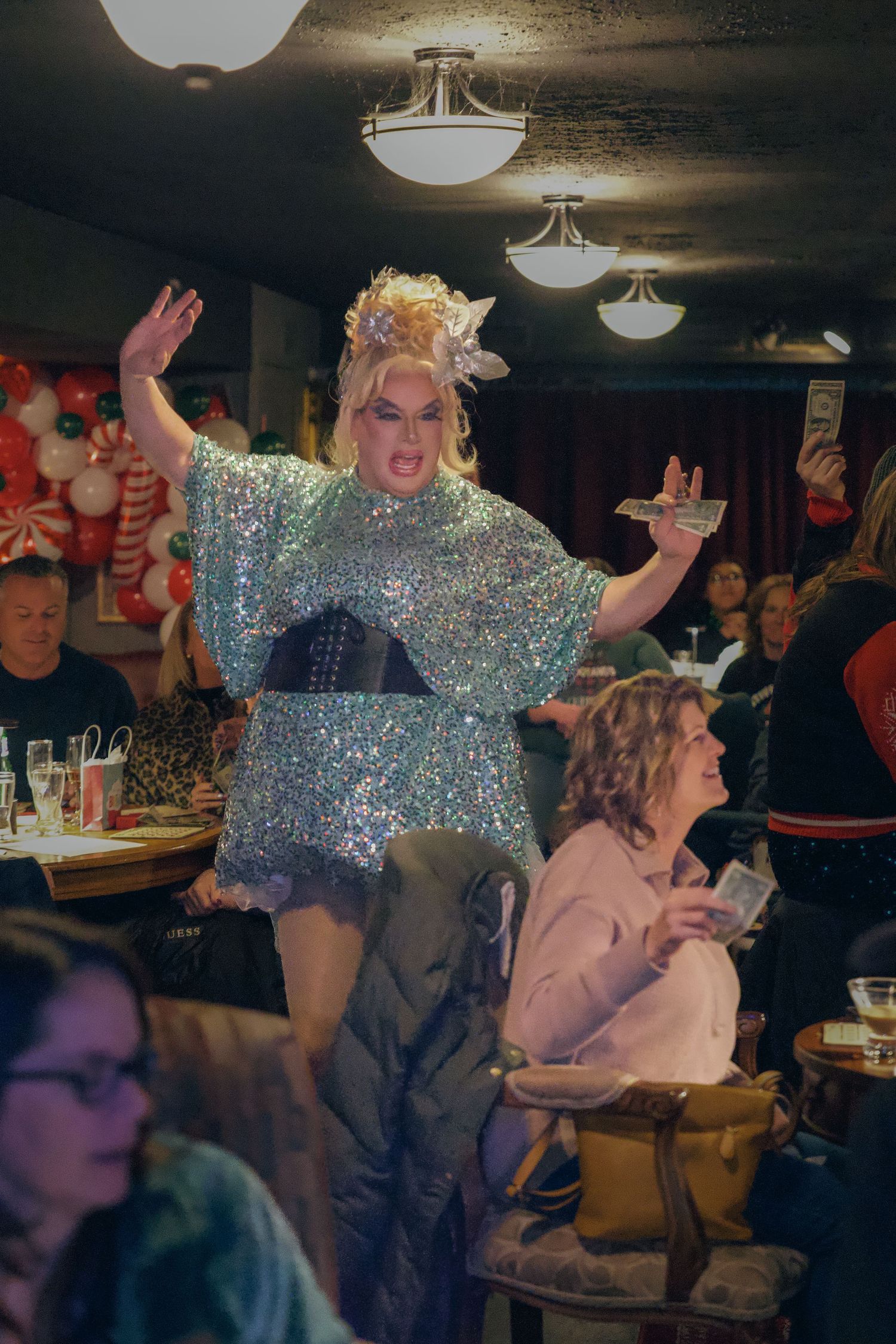 Drag performer in sequined dress entertaining crowd at festive indoor venue.