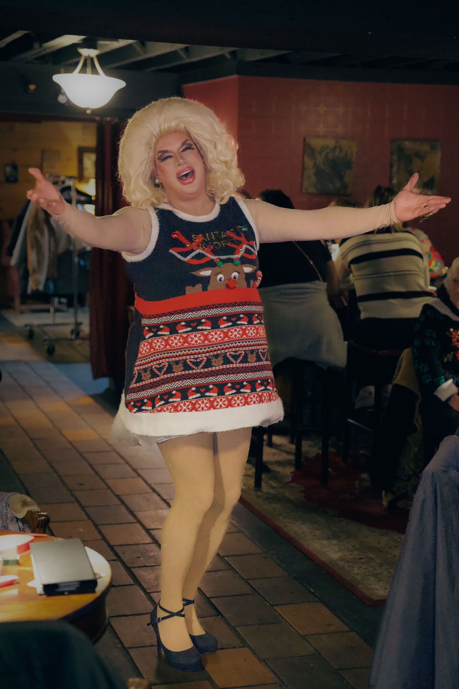 Drag performer in Christmas sweater dress and blonde wig dancing with arms outstretched in a restaurant setting.