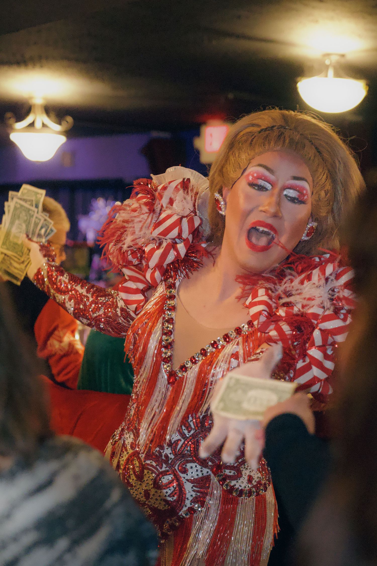 Drag performer in red sequined costume accepting dollar bills from audience at a lively indoor show.