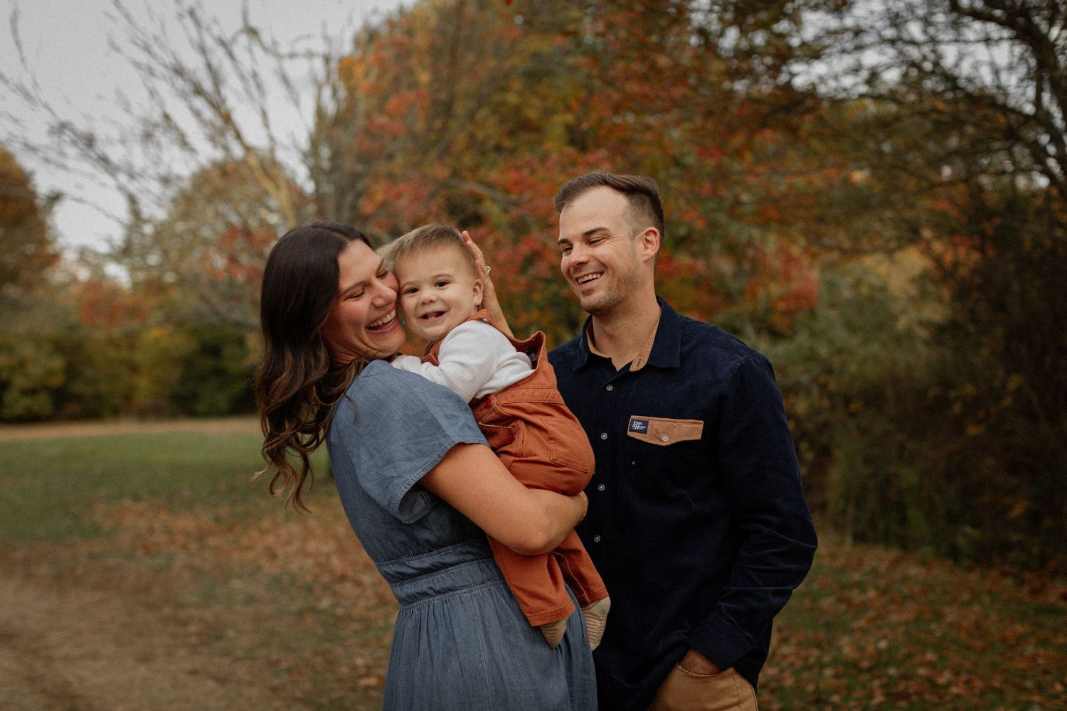 A happy family of three poses outdoors in autumn, parents smiling and holding their toddler among colorful fall foliage.