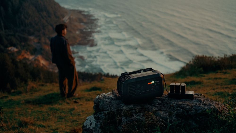 A portable power station sits on a rocky cliff overlooking the ocean at dusk, with a person standing in the background.