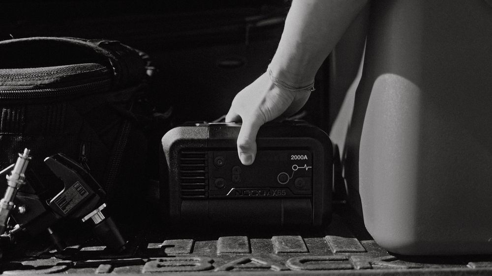 Hand holding a portable 2000A jump starter power bank next to a vehicle in black and white.