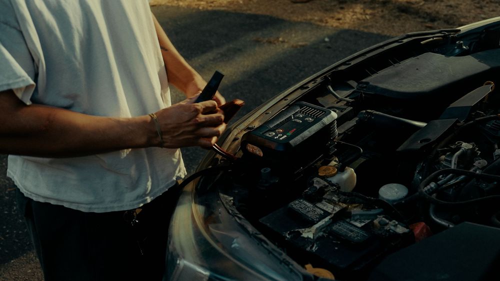 Person checking under open car hood while holding a tool, performing vehicle maintenance outdoors.
