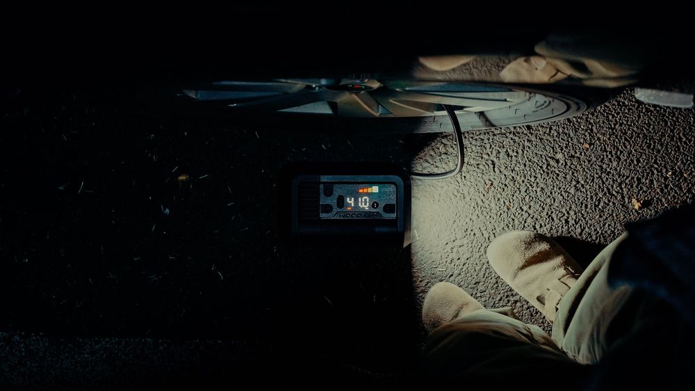 A dimly lit scene showing a smartphone on sandy ground near a person's feet wearing light-colored shoes at night.