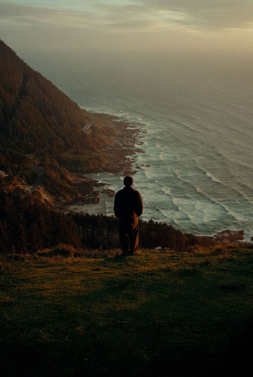 Lone person standing on a grassy coastal cliff overlooking a dramatic ocean shoreline at sunset.