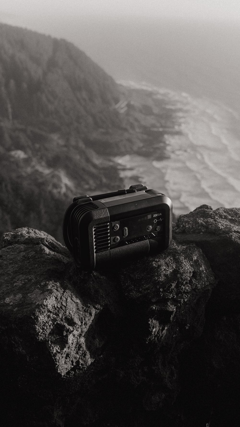 Portable power station placed on rocky mountain cliff with misty coastal landscape in black and white.