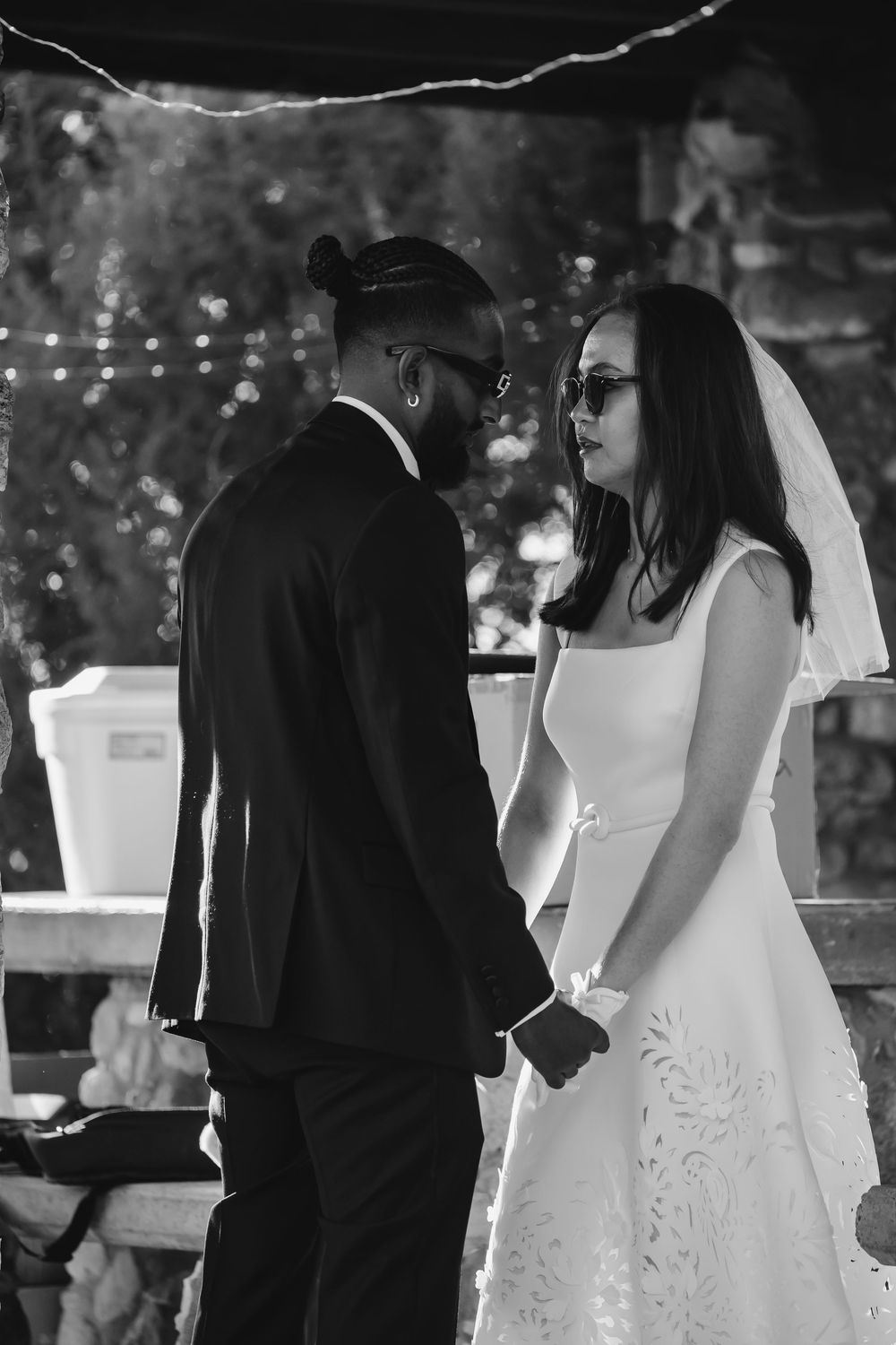 Black and white photo of an interracial couple holding hands at their wedding ceremony, bride in veil and groom in suit.