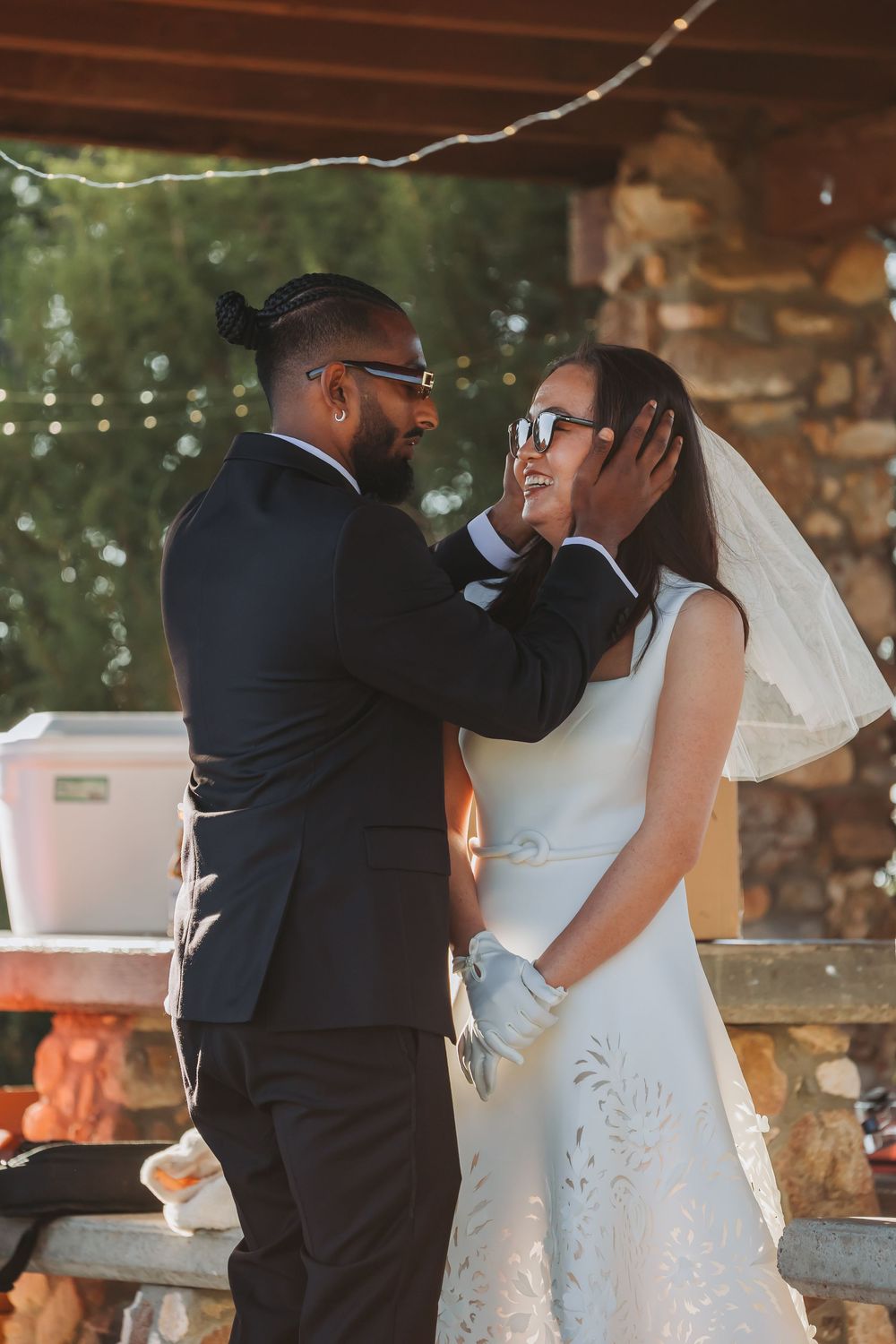 A groom in a black suit tenderly holds his bride's face during an outdoor wedding ceremony, both wearing glasses.