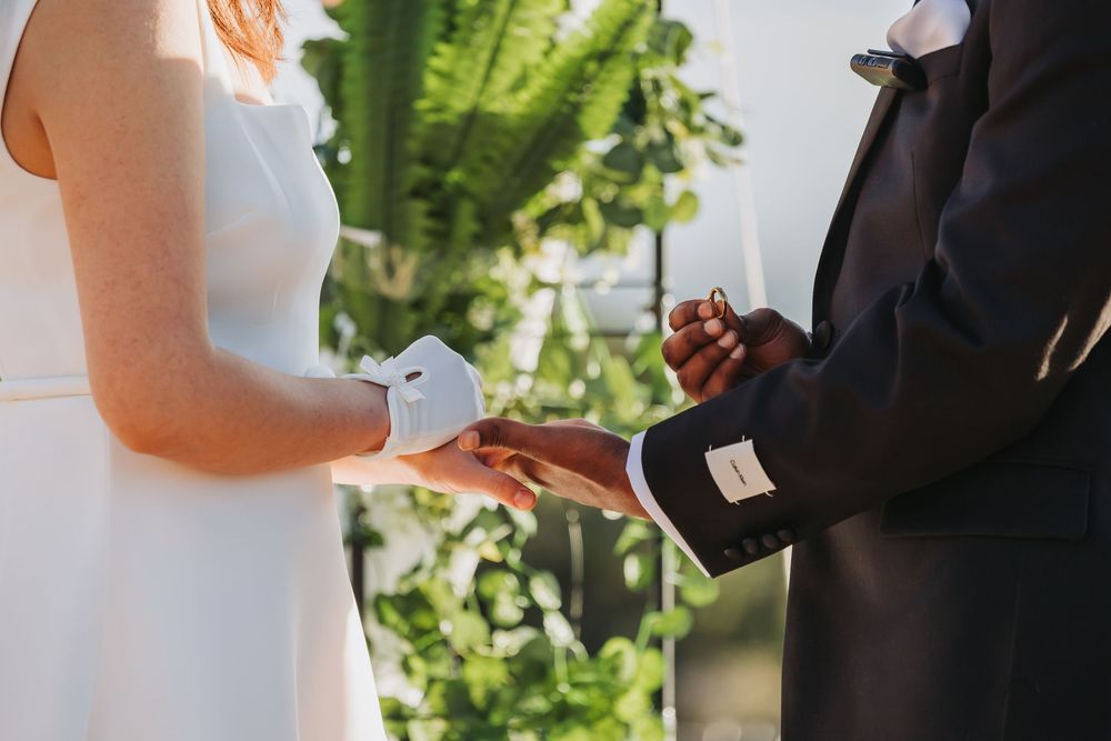 Interracial couple exchanging rings during an outdoor wedding ceremony, bride in white dress and groom in black suit.