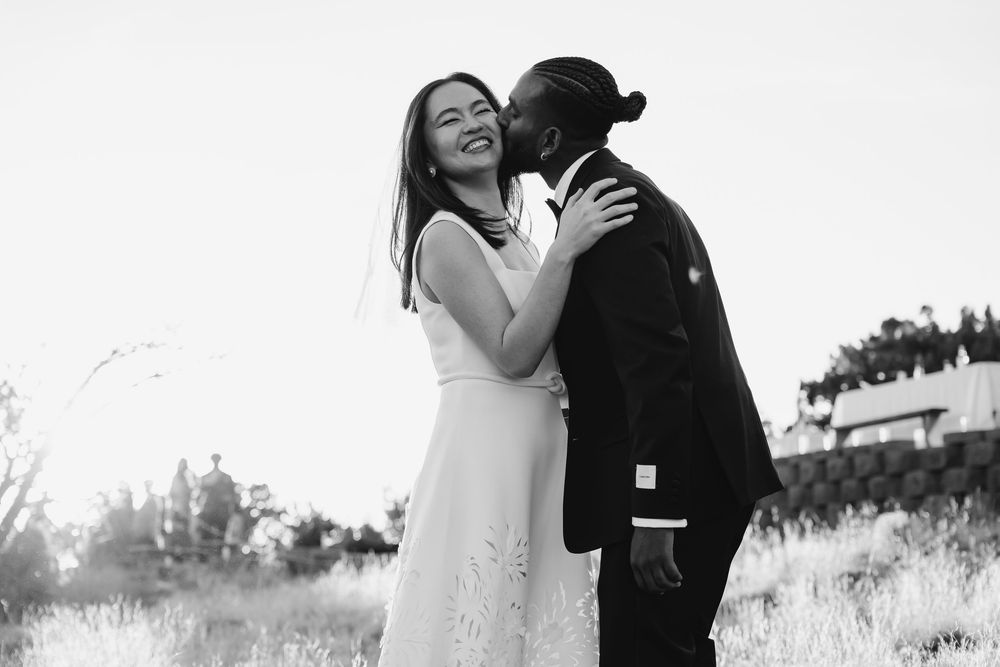 Black and white wedding photo of a joyful couple embracing outdoors, bride in white dress, groom in suit kissing her cheek.