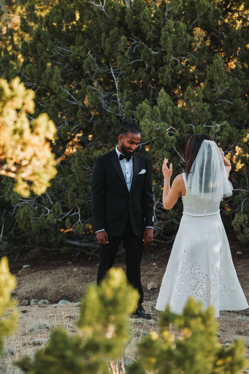 Bride in white lace gown and veil faces groom in black tuxedo during outdoor wedding photo session among evergreen trees.