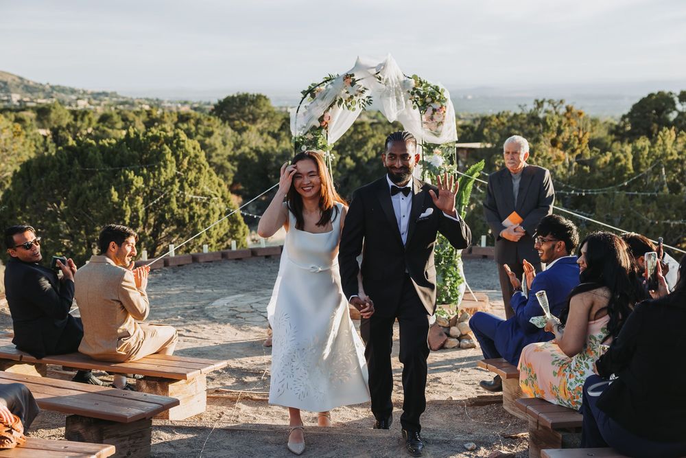 Newlywed couple walks hand-in-hand after outdoor wedding ceremony with floral arch, guests cheering on scenic hilltop venue.