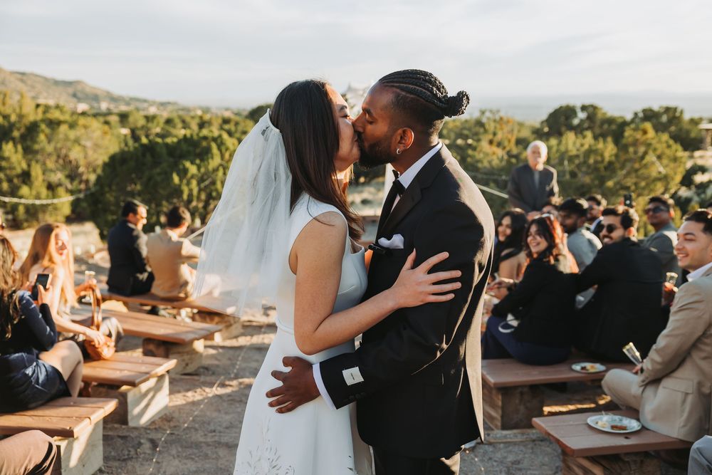 A bride and groom share their first kiss at an outdoor wedding ceremony with guests seated on wooden benches.