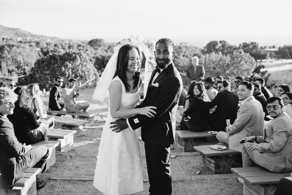 A bride and groom embrace during an outdoor wedding ceremony with guests seated on wooden benches in a scenic hillside setting.