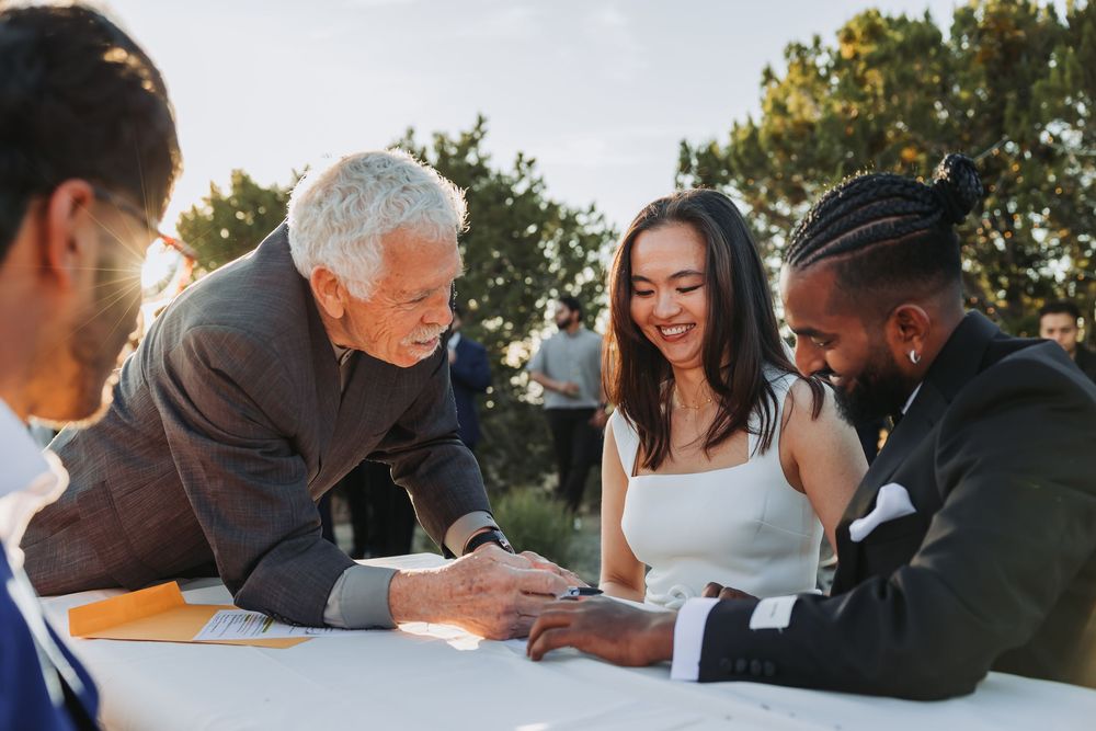 Bride and groom signing wedding documents with an elder officiant outdoors in golden sunlight.