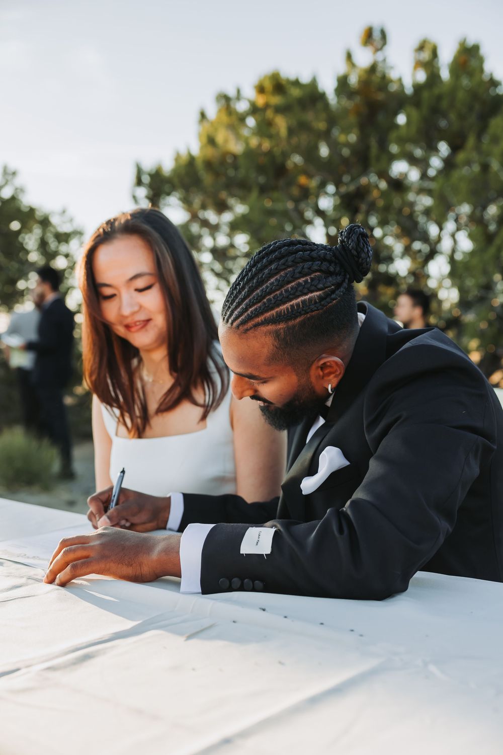Interracial couple signing wedding documents outdoors, groom in black suit with braids, bride in white dress smiling.