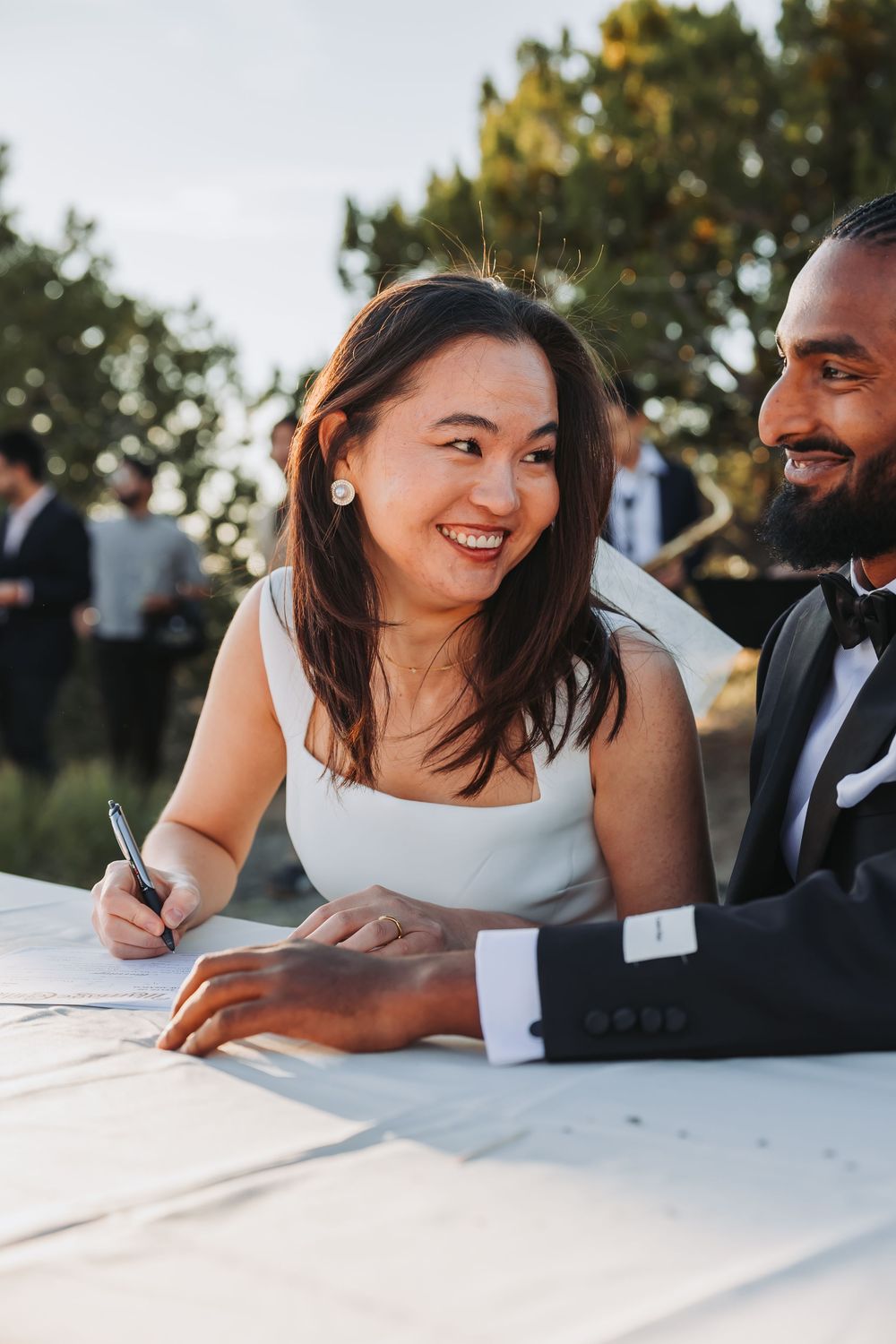 Smiling bride signs marriage certificate beside groom in tuxedo at outdoor wedding table.