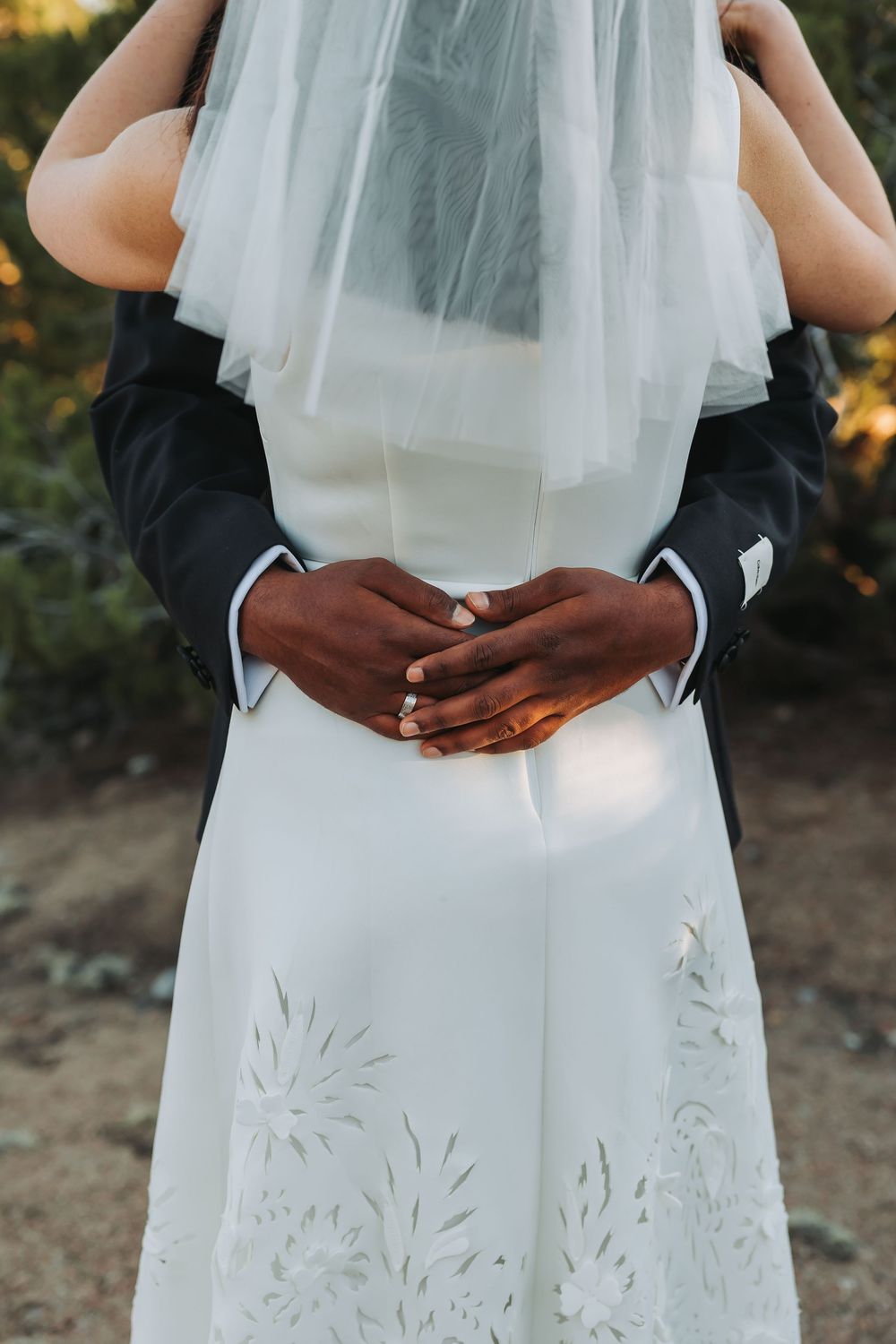 Groom in black suit embracing bride in white floral wedding dress and veil, hands clasped at her back.