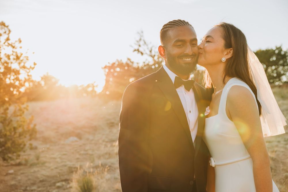 Bride kisses groom on cheek at golden hour sunset, both dressed in elegant wedding attire outdoors.