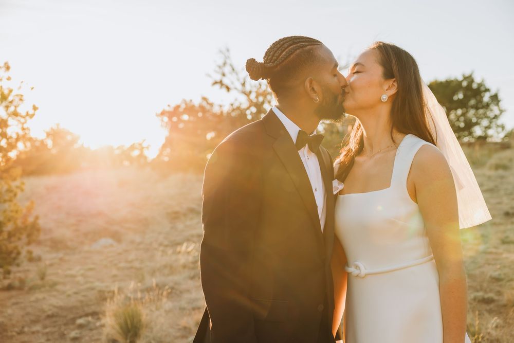 A couple shares a romantic kiss at sunset, bride in white gown with veil, groom in black tuxedo, golden light glowing behind them.
