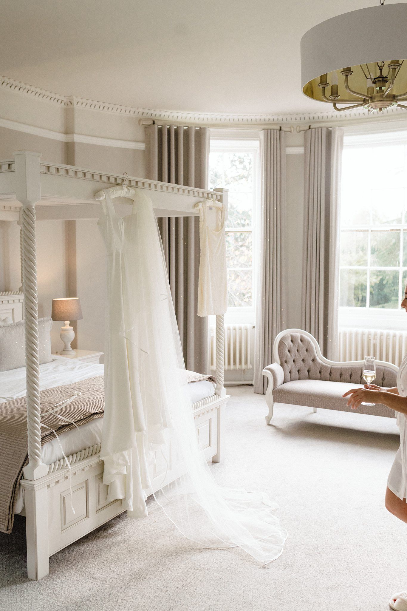 Elegant bridal suite with white four-poster bed, wedding dress hanging, chaise lounge, and bay windows in soft neutral tones.