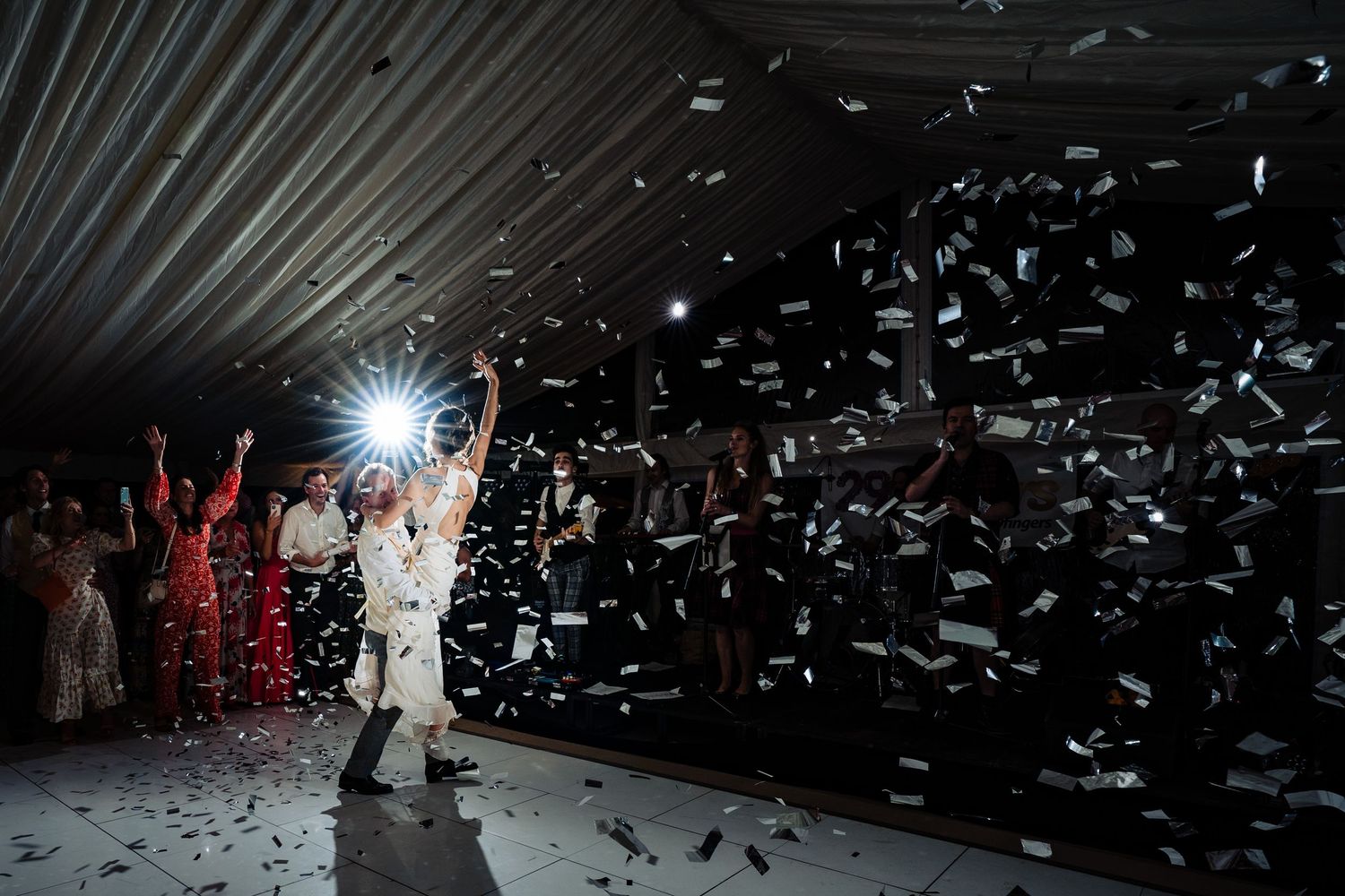Couple celebrates on dance floor with confetti shower and guests cheering in background at festive wedding reception.